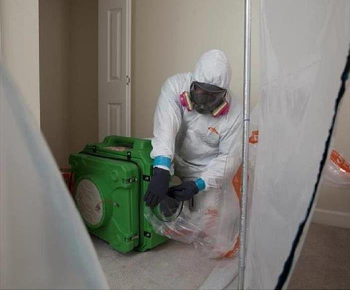 A technician with a biohazard suit and mask setting up plastic barrier around an air scrubber